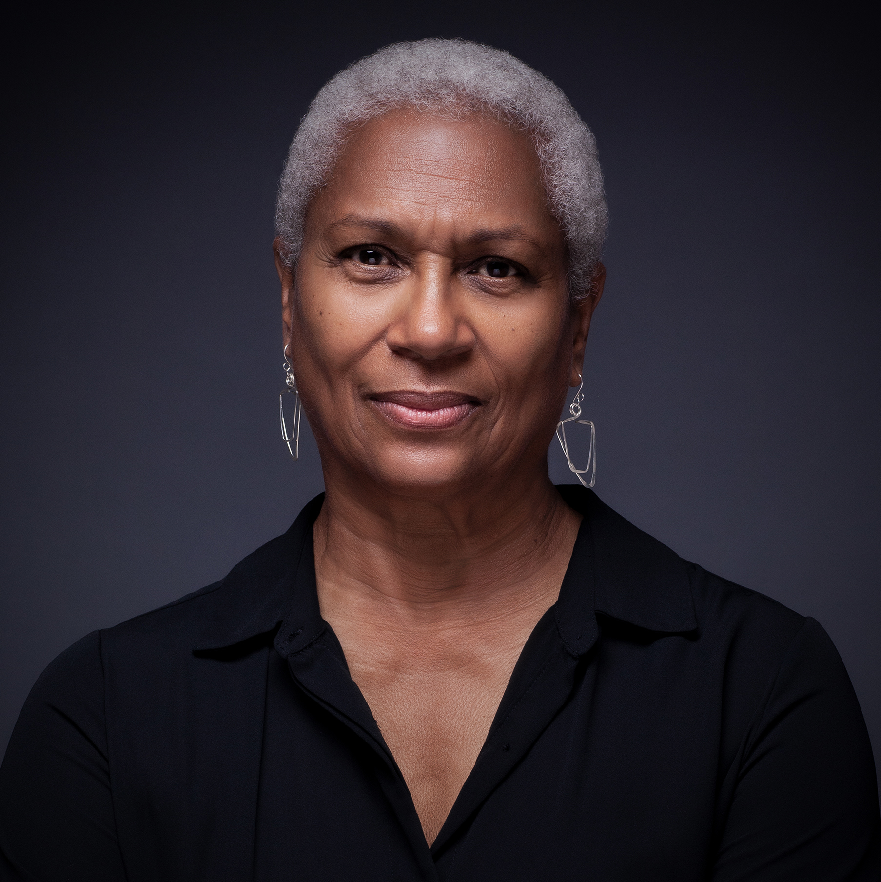 Black woman with white hair facing the camera. She is wearing a black shirt and has silver dangling earrings.